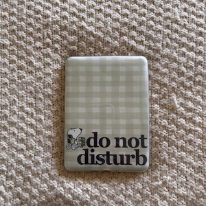 Op de afbeelding: Een lichtgroene en witte telefoonhoes met een ruitpatroon, een cartoon van Snoopy en de woorden "do not disturb" in het zwart. De hoes is rechthoekig en van plastic.