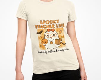 May include: A cream-coloured t-shirt with a Halloween graphic. The design features a cartoon ghost wearing glasses and a bow, holding a book and a pumpkin. Text reads "Spooky Teacher Life" and "Fueled by caffeine & candy corn."