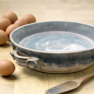 Puede incluir: Una fuente de horno redonda de cerámica con dos asas laterales, con un acabado esmaltado en tonos azul, gris y beige. El recipiente está rodeado por varios huevos marrones y una cuchara de madera, mostrando un aspecto rústico y natural.