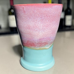 May include: A ceramic tumbler featuring a two-tone glaze. The upper portion displays a soft pink and lavender gradient, while the lower section is a solid light turquoise blue. The vessel has a flared rim and a slightly tapered cylindrical body.