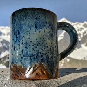 May include: A ceramic mug featuring a speckled blue glaze on the upper portion and a textured, metallic copper-toned base. The mug has a cylindrical body with a curved handle on the right side, showcasing a distinct contrast between the blue and copper.