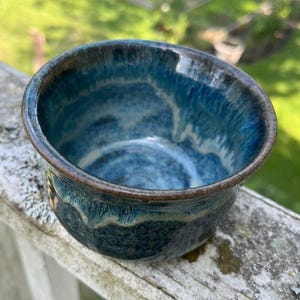 Op de afbeelding: Een handgemaakte keramische kom met een glanzend, blauw glazuur. De kom heeft een afgeronde vorm met een donkerbruine rand en een textuur aan de binnenkant. Het blauwe glazuur heeft variaties van lichte en donkere tinten.