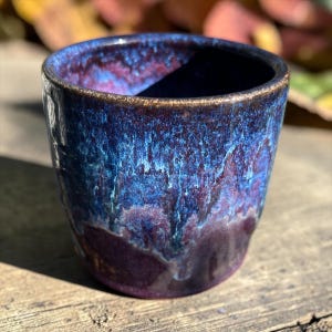 May include: A ceramic cup featuring a deep, glossy glaze with swirling patterns of dark blue, vibrant electric blue, and rich purple. The rim has a textured, metallic-toned finish, and the glaze drips down the sides in a layered effect.