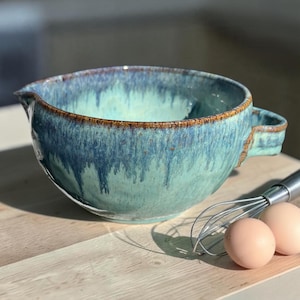 May include: A ceramic mixing bowl with a pour spout and a side handle, featuring a reactive glaze in shades of teal, turquoise, and deep blue with a brown rim. A metal wire whisk and two light brown eggs are placed next to the bowl.