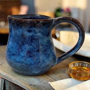 May include: A ceramic mug with a rounded body and a large handle, featuring a deep blue glaze with mottled dark navy and black accents. The surface has a glossy finish with visible variations in color intensity across the curved exterior.