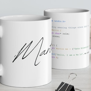 Pode incluir: Uma caneca de cerâmica branca com uma alça preta e o texto "But First, Coffee" em negrito preto. A caneca está sobre um fundo desfocado. A caneca tem aproximadamente 10 cm de altura.