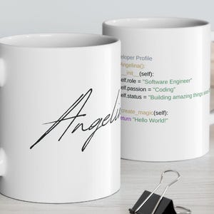 Puede incluir: Dos tazas de cerámica blancas. Una taza tiene el nombre "Angelina" en escritura negra. La otra taza tiene líneas de código con el texto "Software Engineer" y "Coding". Un portátil, clips y una planta están en el fondo.