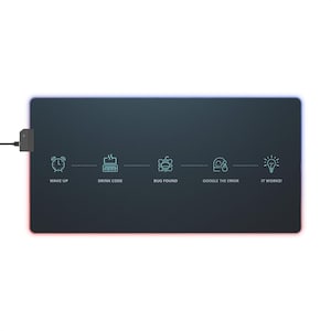 May include: A rectangular black mouse mat with a glowing blue and red edge. The mat features a series of icons and text: "WAKE UP", "DRINK CODE", "BUG FOUND", "GOOGLE THE ERROR", and "IT WORKS!". A small black box is attached to the mat by a wire.