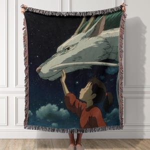 May include: A woven throw blanket featuring a scene from an animated film. The design depicts a young person reaching out to touch a large white dragon against a starry night sky. The blanket has a fringed edge.