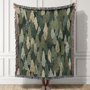 May include: A woven throw blanket featuring a repeating pattern of evergreen trees in various shades of dark green, sage, olive, and cream. The rectangular blanket has a dark background and is finished with a decorative fringed edge on all sides.