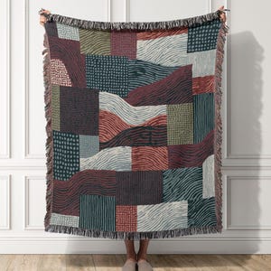 May include: A woven throw blanket featuring a patchwork design of rectangular blocks with various patterns including wavy lines, dots, and vertical stripes in shades of deep red, dark teal, olive green, and cream, finished with a fringed edge.