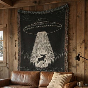 Puede incluir: Una manta tejida en blanco y negro que muestra un gran OVNI sobrevolando un bosque, con un haz de luz que ilumina la silueta de un alce. La manta tiene bordes con flecos y presenta un diseño gráfico de alto contraste.