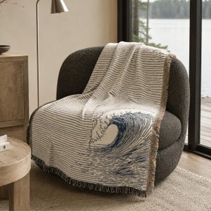 May include: A woven throw blanket featuring a cream-colored base with thin horizontal black lines and a large, detailed navy blue ocean wave graphic. The blanket has a textured appearance with frayed, multi-colored edges draped over a chair.