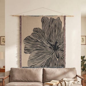 May include: A rectangular woven wall tapestry featuring a large, abstract black floral design on a beige background. The textile has a fringed edge on the sides and is suspended from a horizontal wooden rod with a thin white string.