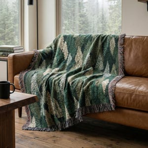 May include: A woven throw blanket featuring a repeating pattern of evergreen trees in shades of dark green, forest green, and cream. The rectangular blanket has a textured finish and decorative fringe along the edges, draped over a brown leather sofa.