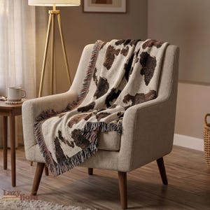 May include: A woven throw blanket featuring a brown and cream cow print pattern with frayed edges. The blanket is draped over a textured cream-colored armchair. The fabric displays a repeating animal hide motif across the entire surface.