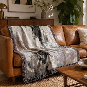 May include: A woven throw blanket featuring an abstract pattern in muted tones of cream, charcoal gray, sage green, and dusty mauve. The rectangular blanket has a textured surface and is finished with decorative fringe along the edges.