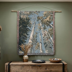 May include: A rectangular woven wall tapestry featuring a low-angle perspective of tall birch trees reaching toward a blue sky. The textile has a fringed edge and is suspended from a wooden rod with a string. The color palette includes shades of blue, white, brown, and tan.