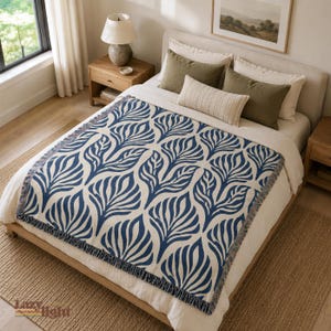 May include: A woven throw blanket featuring a repeating dark blue botanical leaf pattern on a cream background with fringed edges, draped over a bed with white bedding, olive green pillows, and a striped lumbar pillow in a bedroom setting.