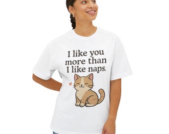 Puede incluir: Camiseta blanca con la frase "I like you more than I like naps". Una ilustración de un gato de dibujos animados está debajo del texto. El gato es marrón y blanco con un corazón rosa. La camiseta la lleva una persona.