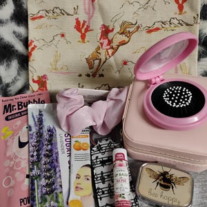 May include: A collection of beauty and self-care items, including a pink compact mirror, lip balm, face masks, and a cosmetic bag with a cowboy and horse print. The items are arranged on a textured surface.