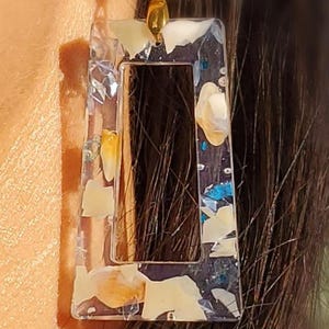 Puede incluir: Pendiente rectangular transparente de resina con herrajes dorados. El pendiente está relleno de fragmentos de concha blancos, amarillos y azules. El pendiente cuelga de un gancho dorado.