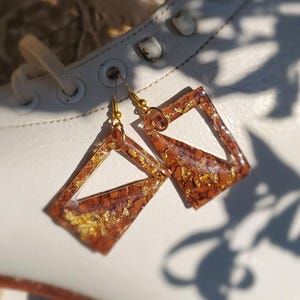 Puede incluir: Un par de pendientes geométricos con forma cuadrada. Los pendientes son una mezcla de marrón y dorado, con escamas doradas incrustadas en la resina. Cuelgan de ganchos dorados. Los pendientes se muestran sobre una superficie blanca.