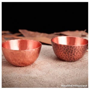 May include: Two small, round copper bowls with a metallic finish. The bowl on the left features a smooth, hammered texture, while the bowl on the right displays a distinct, dense honeycomb-like hammered pattern. The text HealthEnthusiast is visible.