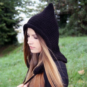 May include: A woman wearing a black knitted hooded hat with a long pointed tip. She is wearing a black dress and has long brown hair.