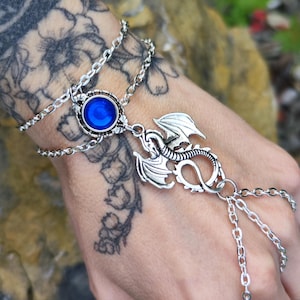 Pu&ograve; includere: Un bracciale a schiava color argento con un cinturino a doppia catena e una pietra cabochon blu circolare al centro. Un ciondolo a forma di drago argentato dettagliato &egrave; fissato al centro, collegato a catene aggiuntive che scendono sulla mano.