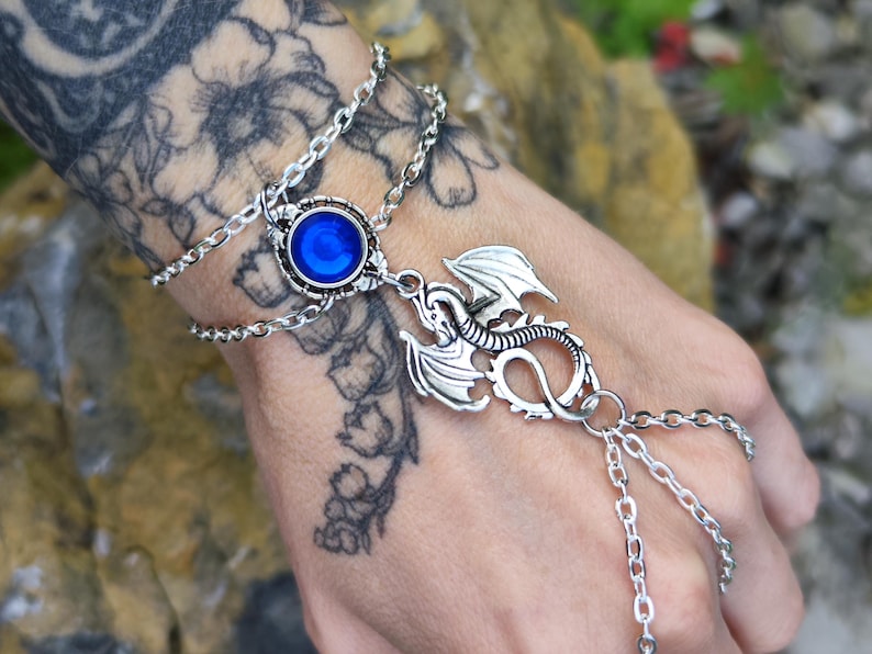 Pu&ograve; includere: Un bracciale a schiava color argento con un cinturino a doppia catena e una pietra cabochon blu circolare al centro. Un ciondolo a forma di drago argentato dettagliato &egrave; fissato al centro, collegato a catene aggiuntive che scendono sulla mano.