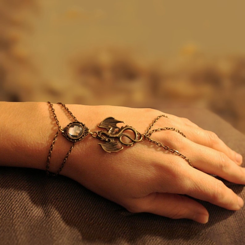 Pu&ograve; includere: Una catena da mano color bronzo con un motivo centrale a forma di drago dalle ali spiegate, collegata a un castone con cristallo trasparente sul bracciale e a delicate catenelle metalliche che avvolgono le dita.