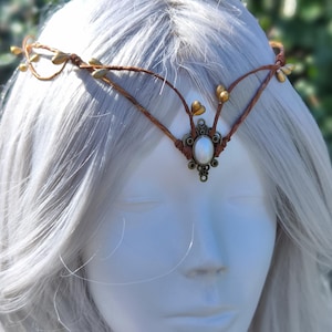 Puede incluir: Una diadema de cuero marr&oacute;n con detalles dorados y una perla blanca en el centro. La diadema est&aacute; puesta en una persona con cabello blanco.