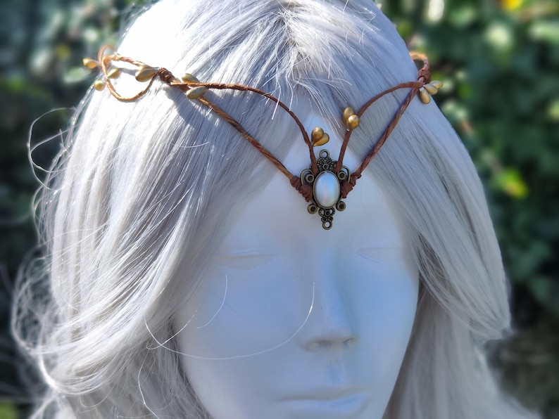 Puede incluir: Una diadema de cuero marr&oacute;n con detalles dorados y una perla blanca en el centro. La diadema est&aacute; puesta en una persona con cabello blanco.
