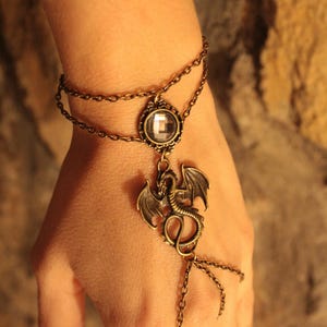 Pu&ograve; includere: Un bracciale schiava color bronzo con catena doppia al polso, un cristallo trasparente sfaccettato circolare centrale e un ciondolo a forma di drago. Il design include una catenina che si estende dal drago a un anello per il dito. Il testo LOS TESOROS DE LA AYALGA &egrave; visibile in alto.