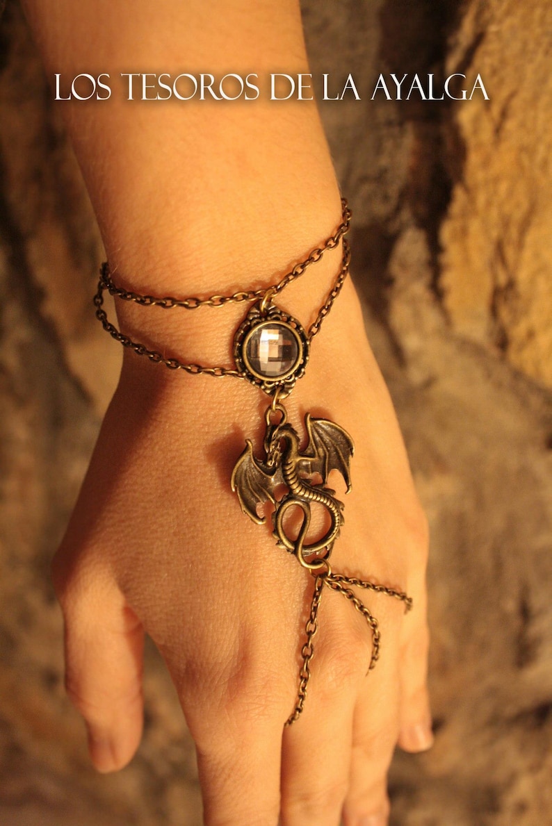 Pu&ograve; includere: Un bracciale schiava color bronzo con catena doppia al polso, un cristallo trasparente sfaccettato circolare centrale e un ciondolo a forma di drago. Il design include una catenina che si estende dal drago a un anello per il dito. Il testo LOS TESOROS DE LA AYALGA &egrave; visibile in alto.
