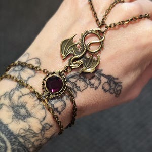 Pu&ograve; includere: Una catena da mano in metallo color bronzo con un ciondolo a forma di drago centrale e una pietra preziosa viola rotonda e sfaccettata. Il gioiello &egrave; composto da delicate catenine che collegano un bracciale a un anello, creando un ornamento.