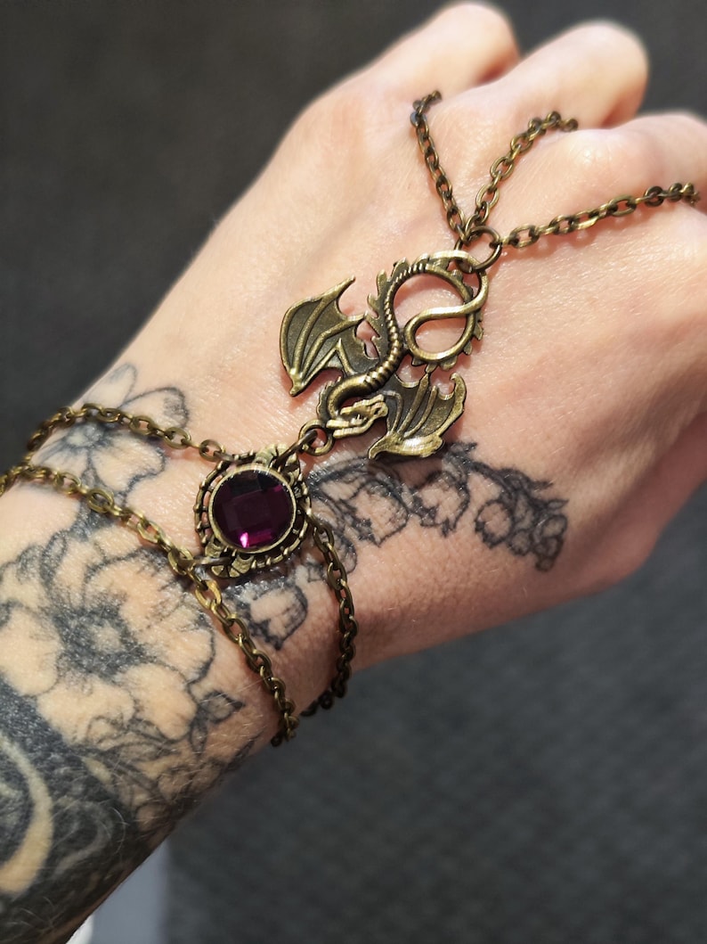 Pu&ograve; includere: Una catena da mano in metallo color bronzo con un ciondolo a forma di drago centrale e una pietra preziosa viola rotonda e sfaccettata. Il gioiello &egrave; composto da delicate catenine che collegano un bracciale a un anello, creando un ornamento.