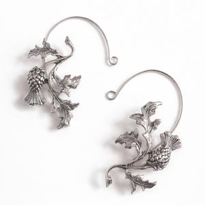 May include: A pair of silver-toned earrings featuring a thistle design. Each earring has a curved wire that hooks through the ear, with a detailed thistle and leaf motif. The earrings are set against a white background.