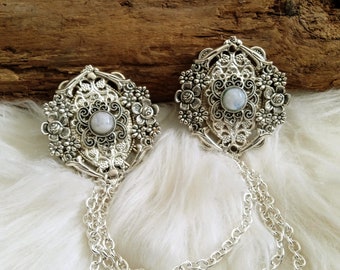 May include: Two silver tone brooches with a white moonstone center and intricate floral designs. Each brooch is attached to a silver chain.