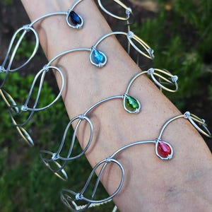 May include: Silver wire bracelets with teardrop-shaped, colorful gemstones. The gemstones are blue, green, and red. The bracelets have a unique, artistic design, with loops and curves. The bracelets are displayed on a person's arm.