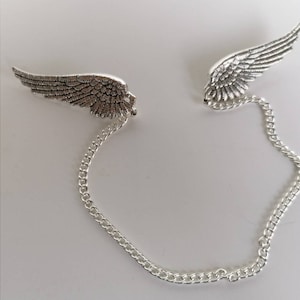 Op de afbeelding: Een zilverkleurige vleugelbroche met gedetailleerde veren, verbonden door een delicate ketting. De broche heeft twee afzonderlijke vleugelontwerpen, elk met ingewikkelde details. De ketting voegt een vleugje elegantie toe aan dit unieke accessoire.