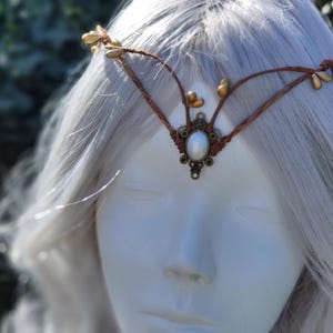 Puede incluir: Una diadema de cuero marr&oacute;n con detalles de hojas doradas y una perla blanca en el centro. La diadema se lleva en una peluca blanca.