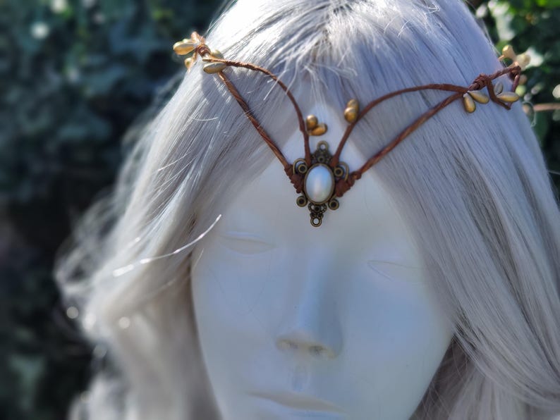 Puede incluir: Una diadema de cuero marr&oacute;n con detalles de hojas doradas y una perla blanca en el centro. La diadema se lleva en una peluca blanca.