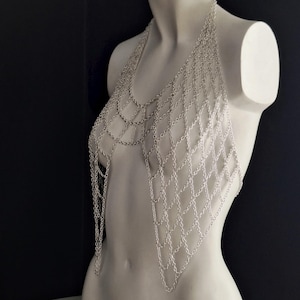 May include: Silver chainmail top with a halter neck design. The chainmail is arranged in a diamond pattern, creating a draped effect. The top is displayed on a white mannequin against a black background.