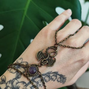 Pu&ograve; includere: Una catena da mano in tonalit&agrave; bronzo antico con un ciondolo a forma di drago centrale e una pietra cabochon rotonda viola. Il gioiello &egrave; composto da delicati anelli in metallo che collegano un bracciale a un anello sul dorso della mano.