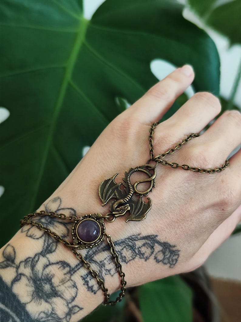 Pu&ograve; includere: Una catena da mano in tonalit&agrave; bronzo antico con un ciondolo a forma di drago centrale e una pietra cabochon rotonda viola. Il gioiello &egrave; composto da delicati anelli in metallo che collegano un bracciale a un anello sul dorso della mano.