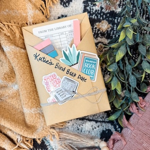 May include: A kraft paper envelope labeled "Katie's Blind Book Date" with stickers and a string tie. The envelope contains a library card, pink pen, and colorful sticky notes. Decorative stickers include a book club design and a potted plant.