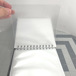 May include: A clear, plastic sheet being held over a white, spiral-bound photo album. The album is open, revealing white pages and clear protective sleeves. The spiral binding is silver.