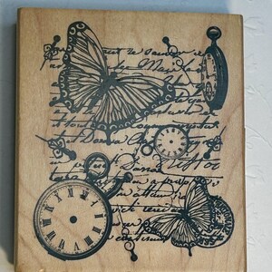 May include: A wooden stamp featuring a butterfly, pocket watches, and script. The butterfly has detailed wing patterns. The pocket watches have Roman numerals. The script is in a cursive font. The stamp is square.